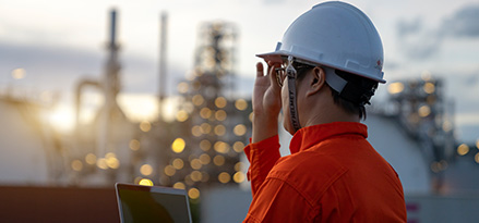 A worker wearing a white hard hat overlooking a power plant as the sun sets behind it