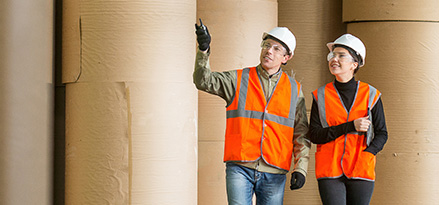 two workers walk alongside each other in a paper mill. One of them is pointing to the paper rolls surrounding them.